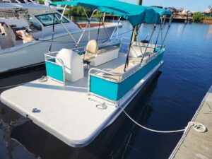 Turquoise pontoon boat docked at a marina with calm water and other boats in the background. - Belle Harbour Marina