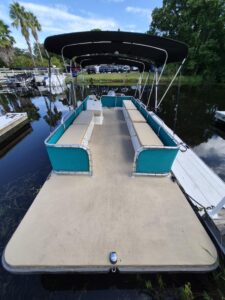 Pontoon boat with teal seating and beige cushions, a large black canopy, docked on calm water. - Belle Harbour Marina