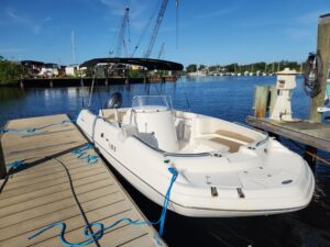 White motorboat moored at a wooden dock with blue lines, calm water, and a marina in the background under a clear blue sky. - Belle Harbour Marina